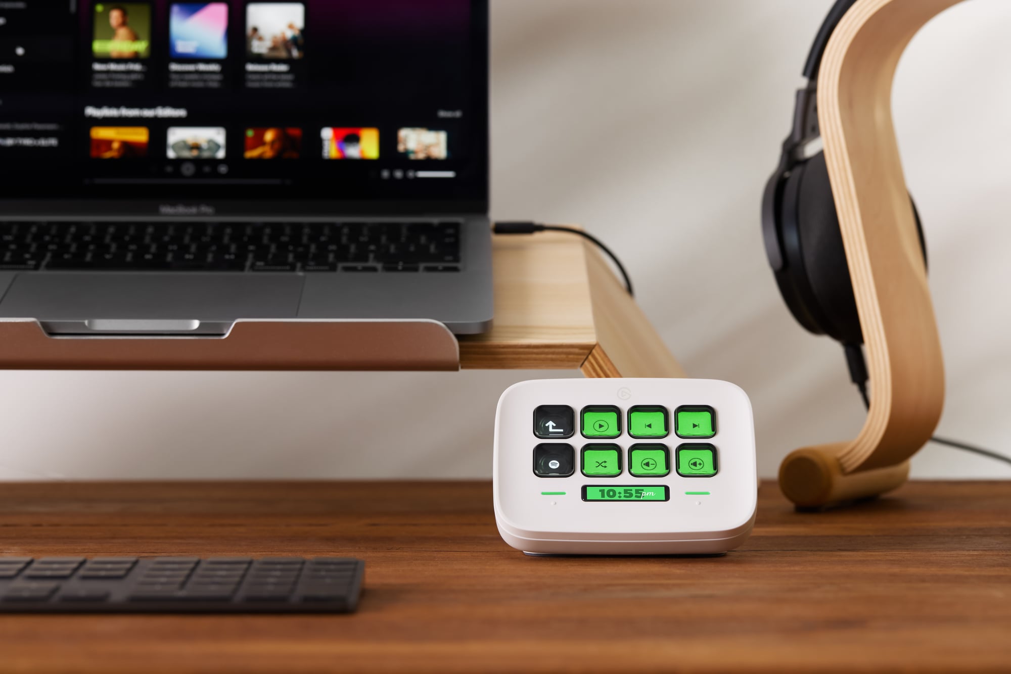 Stream Deck Neo on a wooden desk beside a MacBook showing Spotify — lit keys showing media controls at a glance.